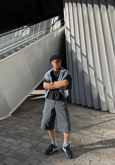 Conjunto casual que incluye una camiseta negra, pantalones cortos de cargo grises y zapatillas negras. Suéter azul drapeado sobre los hombros, gafas de sol redondas.