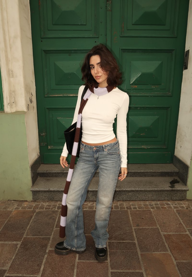 Jovem mulher com cabelo escuro até aos ombros usa uma blusa branca de manga longa, jeans azuis, botas, um cachecol riscado e segura uma bolsa preta, estando à frente de uma porta verde.