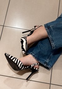 Pieds chaussés de talons hauts noirs et blancs à rayures zébrées, associés à un jean en denim bleu, reposant sur un sol carrelé.