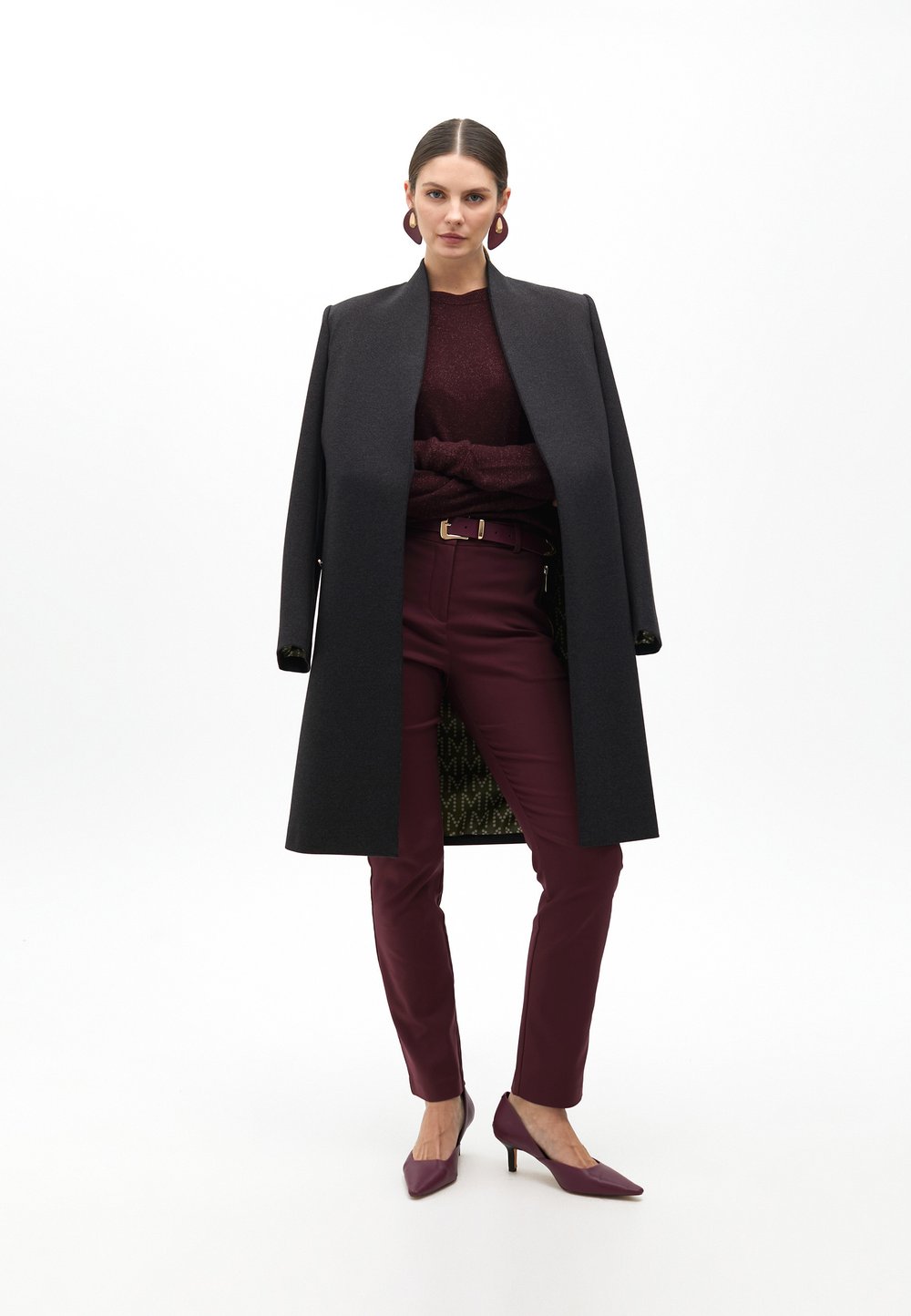 Cappotto lungo nero, maglione bordeaux, pantaloni bordeaux aderenti e tacchi a punta abbinati. Dettagli notevoli: maglione a trama, hardware dorato della cintura, interno patterned.