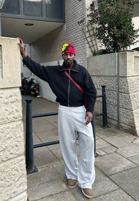 Maglione nero a coste con dettagli zip, pantaloni della tuta grigi chiari e scarpe beige. Berretto colorato con motivi gialli, rossi e neri.