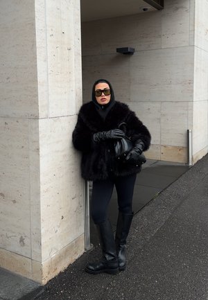 Mujer con abrigo de piel negro, guantes negros, botas y gafas de sol apoyada contra una pared de piedra beige al aire libre.