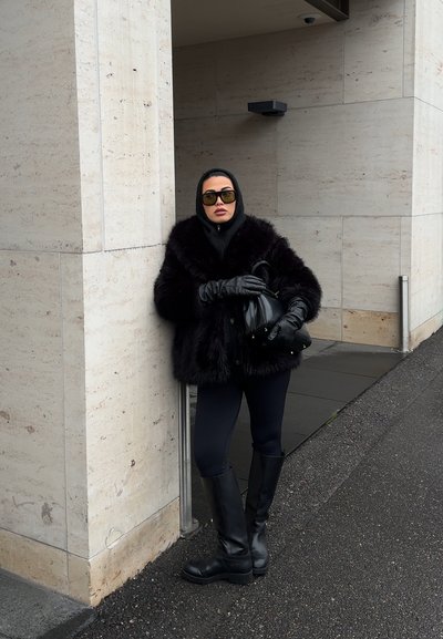Mujer con abrigo de piel negro, guantes negros, botas y gafas de sol apoyada contra una pared de piedra beige al aire libre.