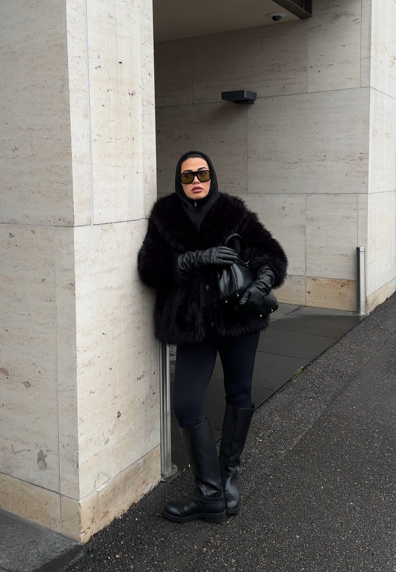 Femme portant un manteau en fourrure noir, des gants noirs, des bottes et des lunettes de soleil, appuyée contre un mur en pierre beige en plein air.