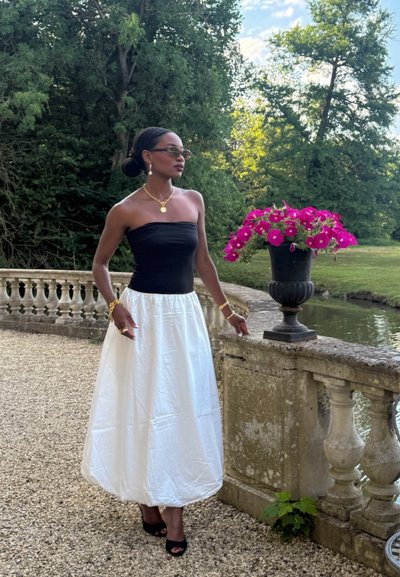 Top negro sin tirantes, falda blanca fluida, combinada con sandalias de tacón negras. Detalles en joyería dorada y gafas de sol. Entorno al aire libre con flores.