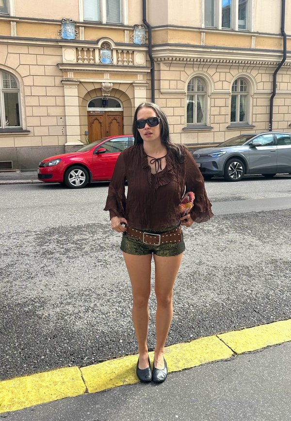 Žena so slnečnými okuliarmi, hnedou blúzkou, zelenými krátkymi nohavicami a čiernymi topánkami stojí na okraji ulice s budovami a zaparkovanými autami za ňou.