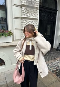 Beige oversized yllekappa, brun och cream stickad tröja, svarta byxor och en rosa vävd väska; utomhus i en urban miljö med kullerstensmark.