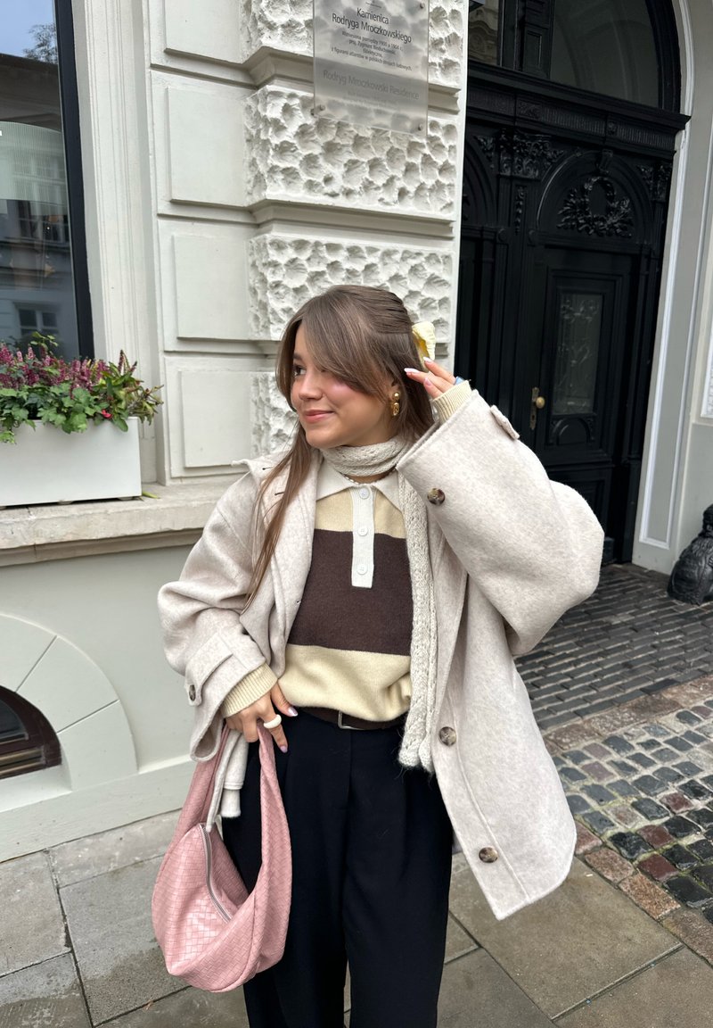 Beige oversized ullfrakk, brun og kremfarget strikkegenser, svarte bukser og en rosa vevd veske; utendørs urbant miljø med brosteinsbelagt underlag.