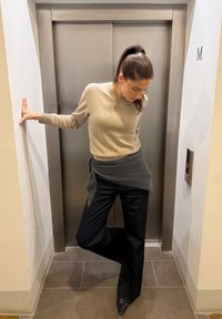 Beige ulltröja, mörkgrå draperad tyg vid midjan, svarta vida byxor och spetsiga svarta klackskor nära en hiss i neutrala omgivningar.