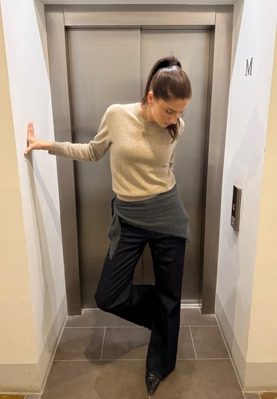 Suéter de lana beige, tela gris oscuro drapeada en la cintura, pantalones negros de pierna ancha y tacones puntiagudos negros cerca de un ascensor en un entorno neutro.
