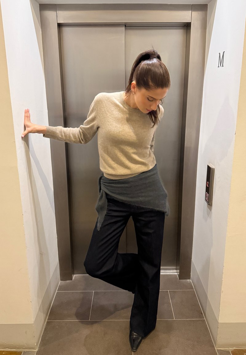 Camisola de lã bege, tecido cinzento escuro drapeado na cintura, calças pretas de perna larga e saltos pontiagudos pretos junto a um elevador em ambiente neutro.