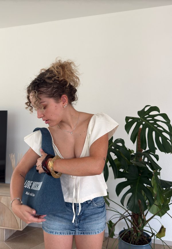 En ung kvinde justerer sin blå Love Moschino tote-taske, mens hun står indendørs nær en stor potteplante af Monstera.