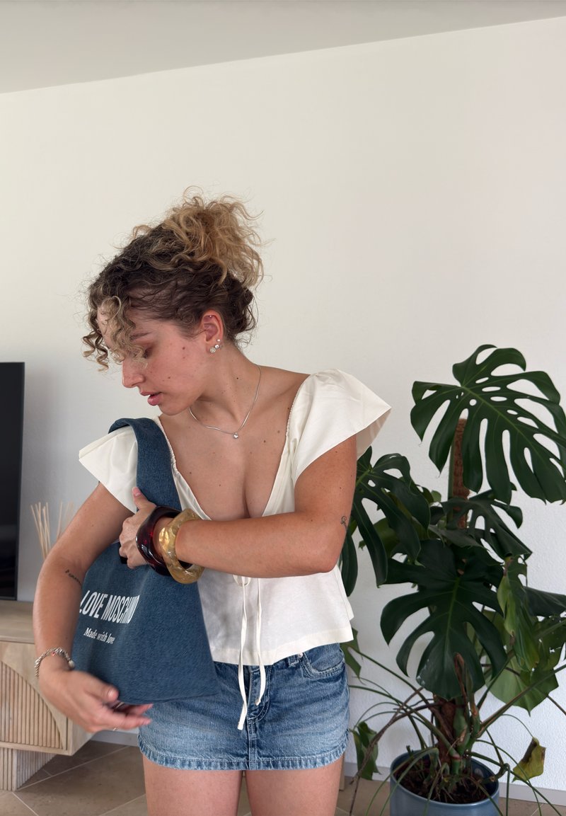 Jeune femme ajustant son sac fourre-tout bleu Love Moschino tout en se tenant à l'intérieur près d'une grande plante Monstera en pot.