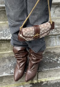 Botas marrones de cuero hasta la rodilla con punta puntiaguda, combinadas con un bolso de mano con estampado marrón y beige que incluye una correa de cadena dorada y detalles de hebilla.
