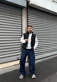 Must tikitud vest valge pika varrukaga särgi peal, kombineeritud laia säärega tume teksapükstega ja hallide tossudega linnahõngulises keskkonnas.