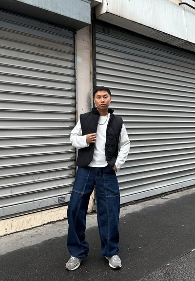 Chaleco negro acolchado sobre una camisa blanca de manga larga, combinado con vaqueros de mezclilla oscuros de pierna ancha y zapatillas grises en un entorno urbano.