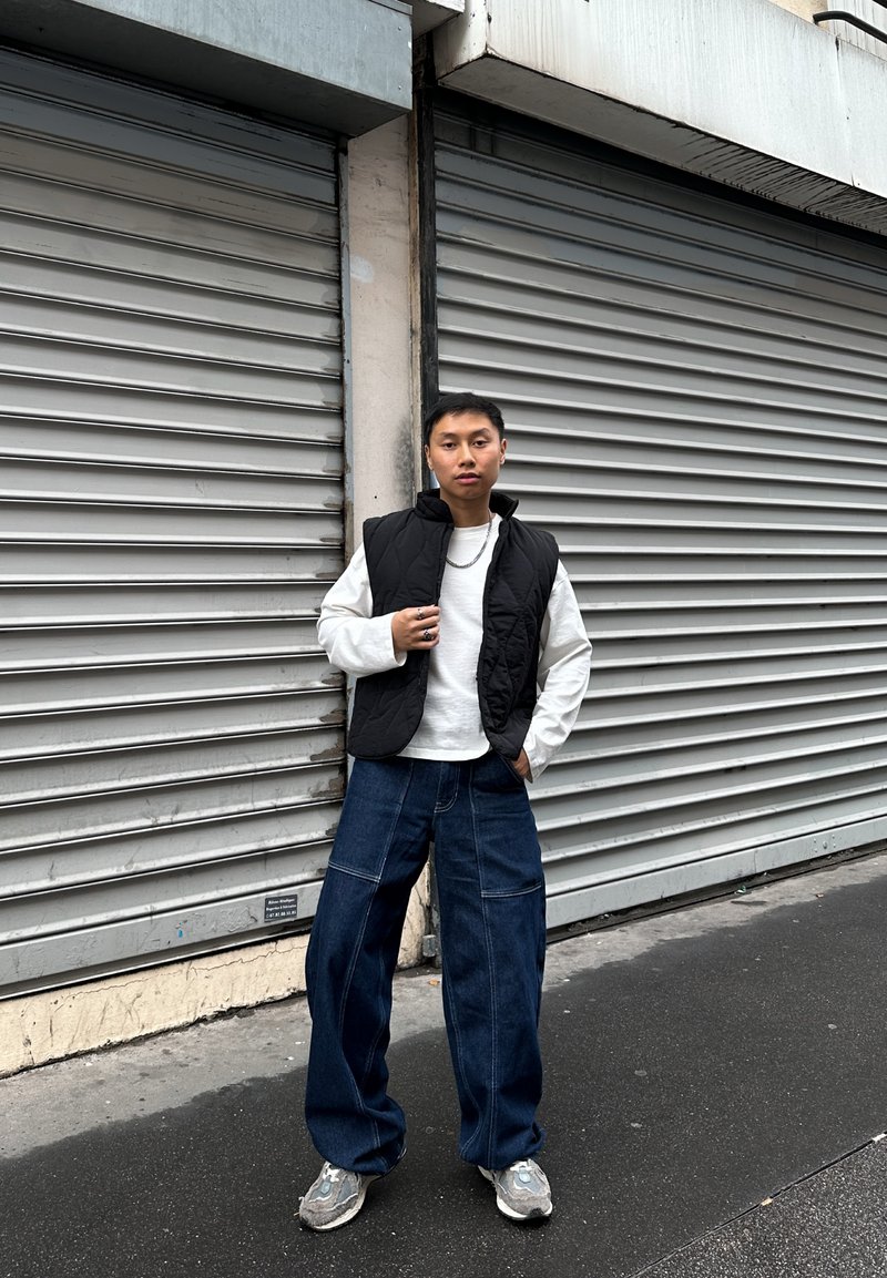 Colete preto acolchoado por cima de uma camisa branca de manga comprida, combinado com jeans escuros de perna larga e sapatilhas cinzentas num ambiente urbano.