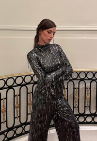 Femme aux cheveux foncés, vêtue d'une tenue noire à col haut et manches longues, ornée de rayures verticales brillantes, se tenant les bras croisés près d'une balustrade décorative.