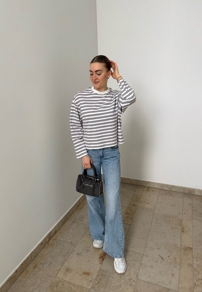 Mujer con una camisa de manga larga a rayas, vaqueros de pierna ancha azul claro, zapatillas blancas, sosteniendo un pequeño bolso negro, de pie en el interior.