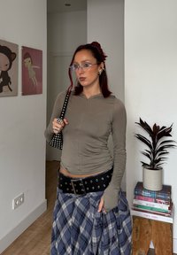 Top beige a maniche lunghe con zip, borsa nera con borchie, cintura nera con occhielli e gonna a quadri blu. Occhiali trasparenti e gioielli argentati.