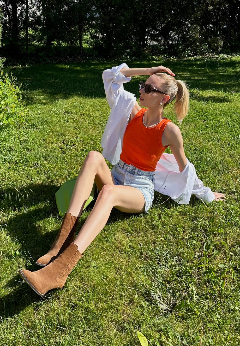 Camisola laranja, camisa oversized branca, calções de ganga, botas de camurça castanhas até ao tornozelo com costura vertical, sentada na relva com uma bolsa verde.
