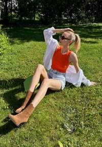Camisola laranja, camisa oversized branca, calções de ganga, botas de camurça castanhas até ao tornozelo com costura vertical, sentada na relva com uma bolsa verde.