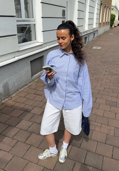 Camisa de rayas azul y blanca de gran tamaño, pantalones cortos blancos, zapatillas plateadas, sosteniendo un teléfono, de pie sobre una acera de ladrillos.