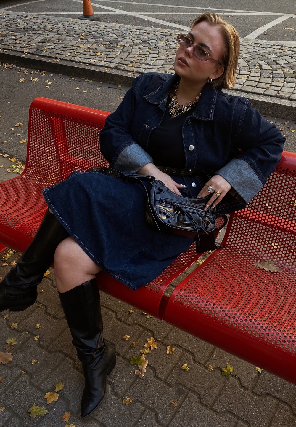 Τζάκετ τζιν με ανοιχτό μπλε μανίκια, μαύρη μπλούζα και σκούρα τζιν φούστα. Φορεμένο με μαύρες μπότες μέχρι το γόνατο και μικρή μαύρη τσάντα. Σε μια κόκκινη πάγκο.