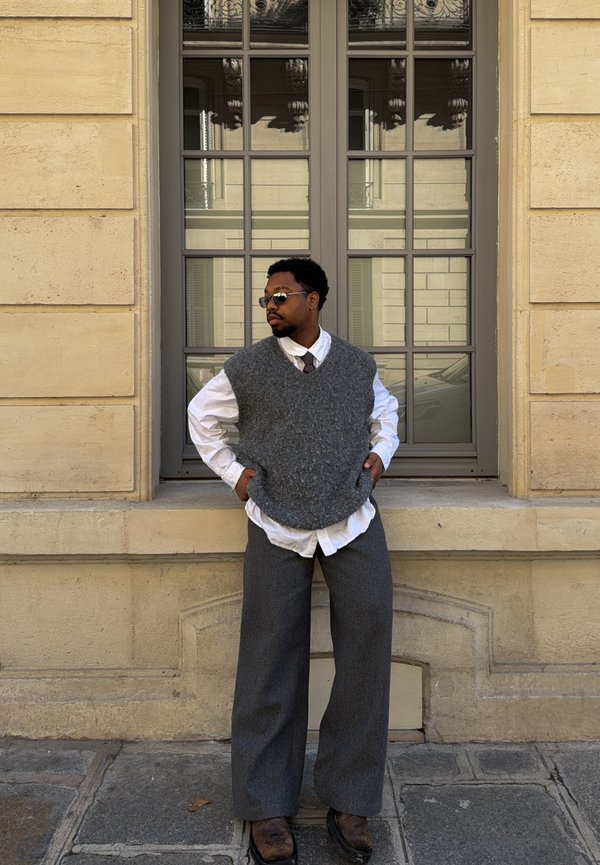 Mees, kes kannab halli vestit, valget särki, lipsu ja päikeseprille, seisab käed taskutes beeži seina ja akna ees.