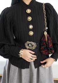 Blouse noire plissée avec boutons dorés et en perle, cintrée à la taille par une ceinture noire avec une boucle ornée. Jupe grise, sac à main texturé.