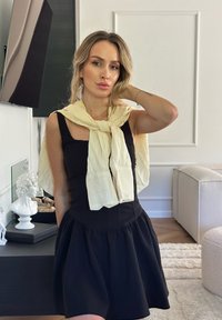 Vestido preto sem mangas com saia evasê, combinado com um lenço listrado em amarelo claro amarrado ao pescoço. Fundo neutro com elementos decorativos.