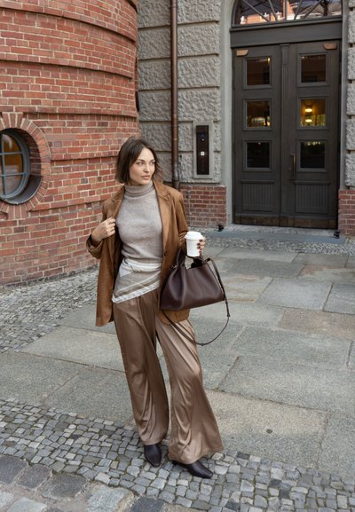 Blazer de ante marrón sobre un cuello de tortuga beige, combinado con pantalones fluidos de un marrón claro. Un bolso de mano marrón oscuro y una taza de café completan el look.