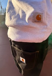 Grauer Sweatshirt mit gerippter Textur, mit einem quadratischen Carhartt-Label. Schwarze Hose mit gelben Nähten und einem Carhartt-Tag auf der Gesäßtasche.