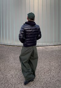 Homme portant une doudoune noire, une casquette verte et un pantalon vert ample, debout les mains dans les poches, dos tourné vers la caméra en plein air.