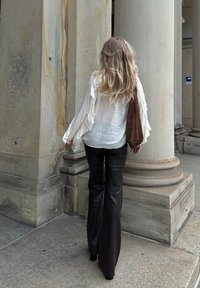 Witte, gerimpelde blouse met lange mouwen, gecombineerd met hoge, uitlopende zwarte leren broek; model met een bruine suède tas, lopend langs kolommen.