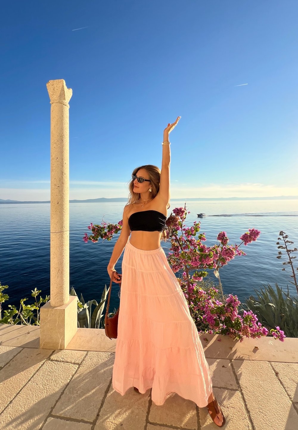 Weißer, gestufter Maxirock mit strukturierter Oberfläche und schwarzem trägerlosem Oberteil. Der Hintergrund zeigt einen Steinsäule und lebendige rosa Blumen am Meer.