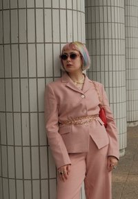 Rosa dress med en knappet blazer og vide bukser, parret med et kædebælte, perlekæde og store solbriller. Tekstureret baggrund.