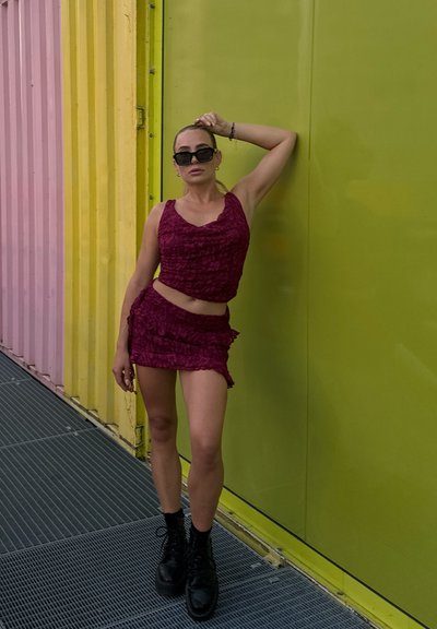 Conjunto de top corto texturizado en color marrón y minifalda, adornado con bordes acampanados. Botas negras con cordones. Fondo en verde brillante y rosa pastel.