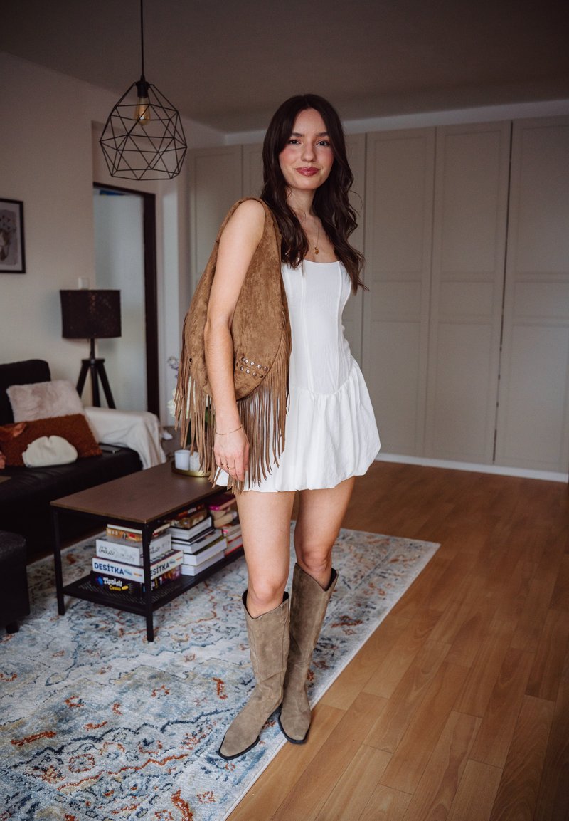 Jeune femme en robe blanche portant des bottes en daim couleur beige et un sac à franges, debout dans un salon avec un tapis à motifs et des livres sur une table basse.