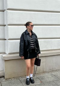 Czarna skórzana kurtka, w paski top, czarna mini spódniczka, białe skarpetki i czarne buty. Akcesorium: mała torebka. Neutralne tło.