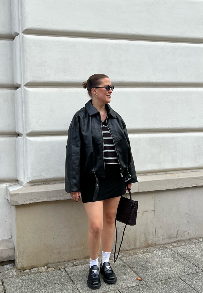 Veste en cuir noire, haut rayé, mini-jupe noire, chaussettes blanches et chaussures noires. Accessoire : petit sac à main. Fond neutre.