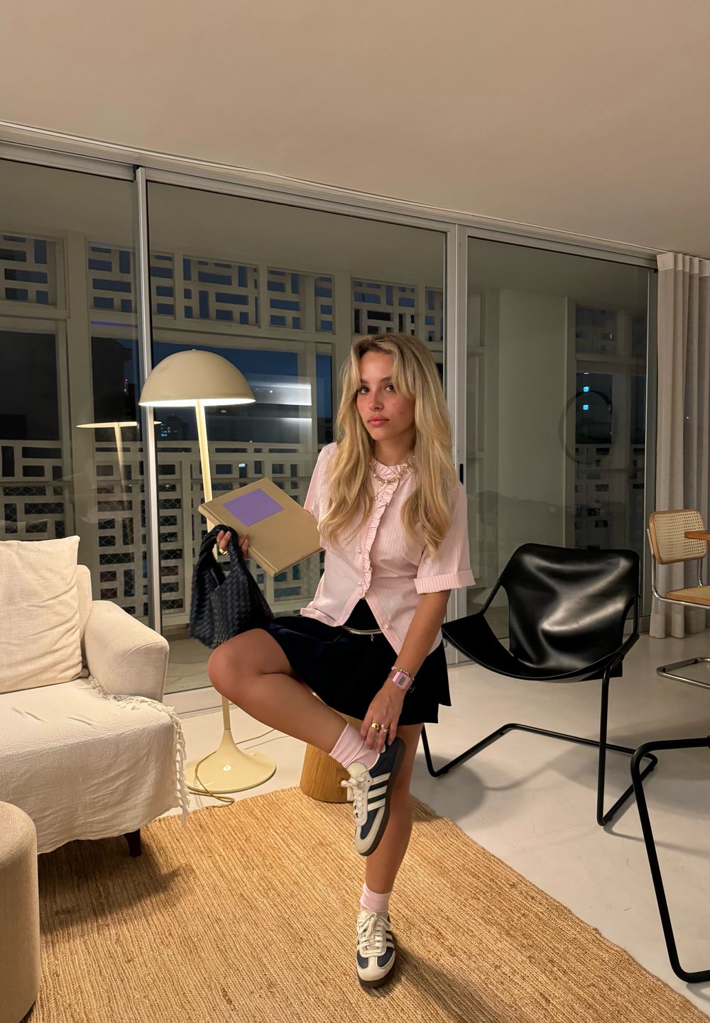 Helles rosa kurzärmliges Blusenshirt mit Rüschen, schwarzer Rock, weiße und marineblaue Sneakers, hält eine Mappe, sitzt in einem modernen Raum.