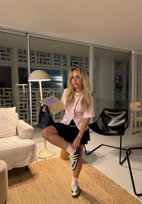 Helles rosa kurzärmliges Blusenshirt mit Rüschen, schwarzer Rock, weiße und marineblaue Sneakers, hält eine Mappe, sitzt in einem modernen Raum.