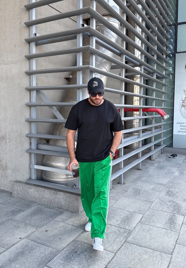 Czarny t-shirt zestawiony z zielonymi spodniami sportowymi z białymi paskami po bokach; białe sneakersy i okulary przeciwsłoneczne; trzymając napój.