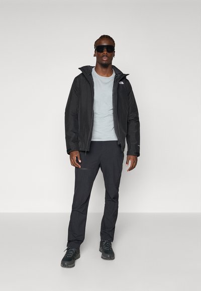 Chaqueta negra con capucha sobre una camiseta gris claro, combinada con pantalones oscuros y zapatos negros. El atuendo presenta un diseño elegante y materiales prácticos.