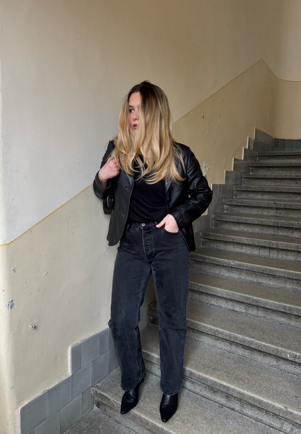 Femme aux longs cheveux blonds portant une veste en cuir noire, un t-shirt noir, un jean noir et des bottes noires, debout sur un escalier en pierre à l'intérieur.