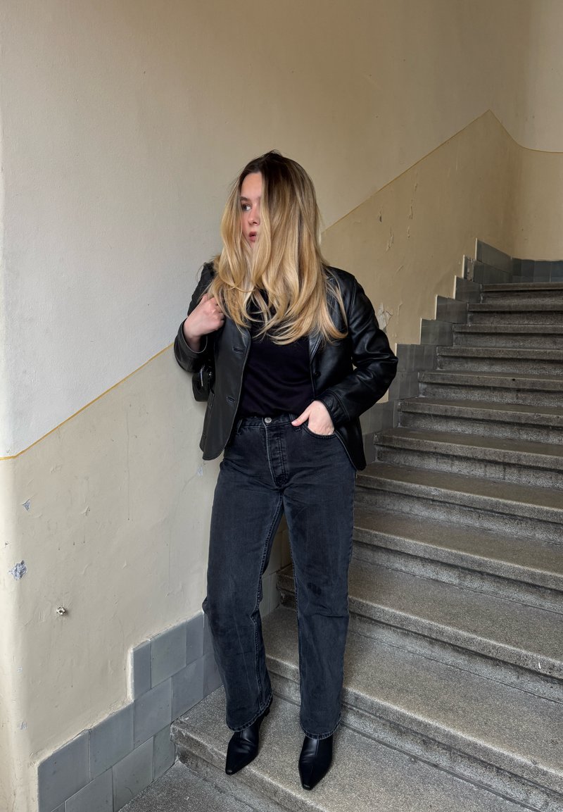 Femme aux longs cheveux blonds portant une veste en cuir noire, un t-shirt noir, un jean noir et des bottes noires, debout sur un escalier en pierre à l'intérieur.