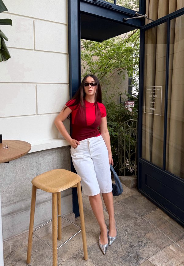 Frau mit Sonnenbrille, trägt ein rotes Top, weiße knielange Shorts und silberne Absätze, steht neben einem Holzschemel nahe einer offenen Tür mit Grün draußen.