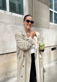 Femeie îmbrăcată într-o haină de trench bej și ochelari de soare negri stă în aer liber ținând un pahar cu smoothie verde pe care scrie "Encré Café".