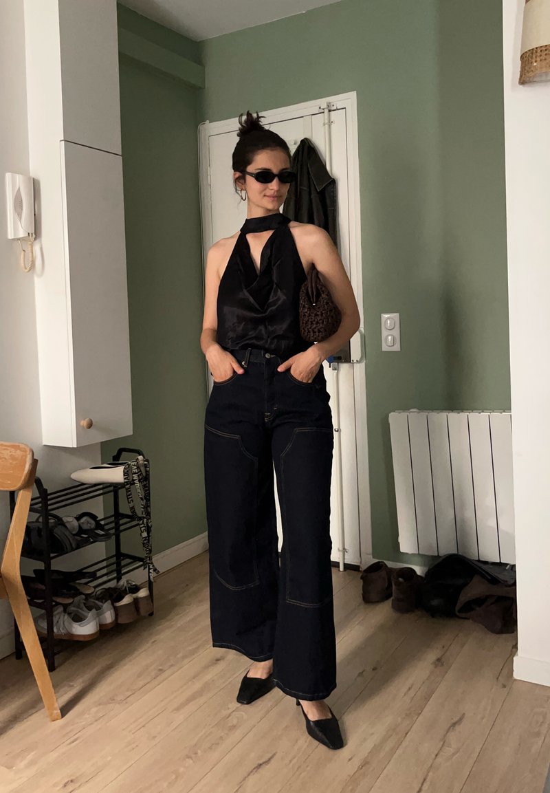 Femme portant un haut dos nu noir, un jean foncé à jambes larges, des chaussures noires, des lunettes de soleil et tenant un sac à main marron dans une pièce aux murs verts.